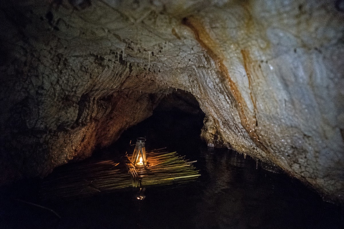 A raft of flax stems floating on black water at the mouth of a low limestone cave with a candle lantern burning on it