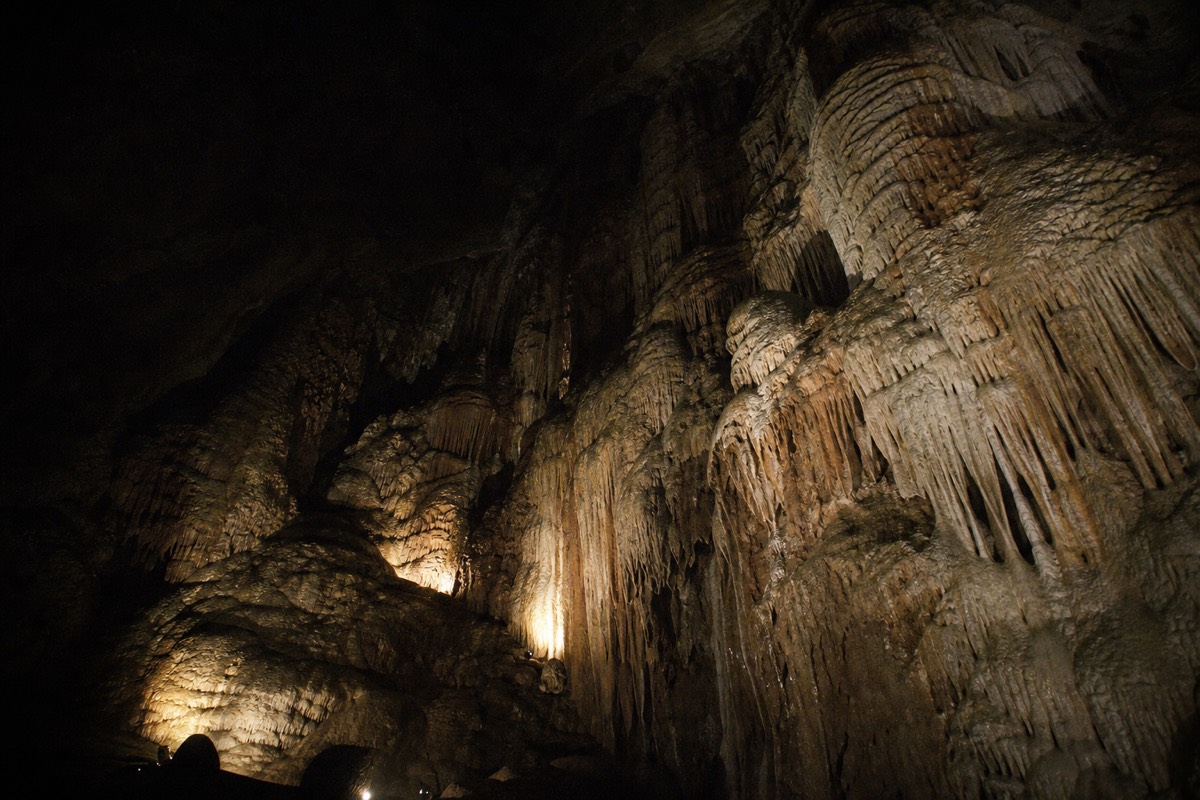 A vast domed cave chamber with layered flowstone walls lit only by weak warm uplights on the floor and a ceiling vanishing into black