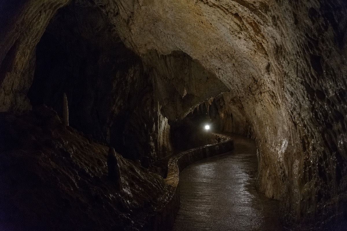 An empty formed walkway inside a limestone cave curving out of sight under one weak lamp with absolute dark beyond