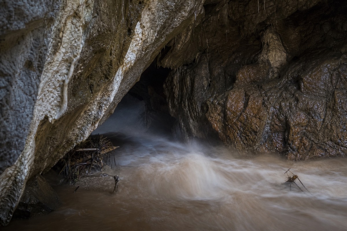 A brown swollen river squeezing into a limestone cave mouth after heavy rain with debris caught on a rock lip